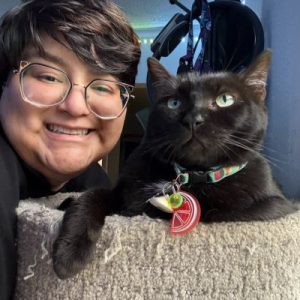 Smiling pet sitter with short hair and glasses posing beside a relaxed black cat with green eyes