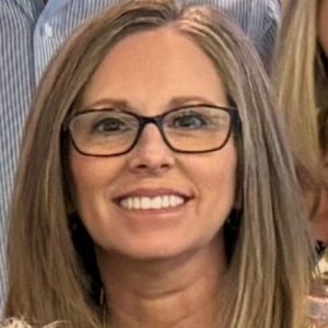 Smiling woman with straight, shoulder-length light brown hair wearing glasses and a light-colored top
