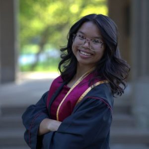 A young woman wearing graduation robes and a maroon stole with gold trim stands outdoors, smiling confidently with her arms crossed